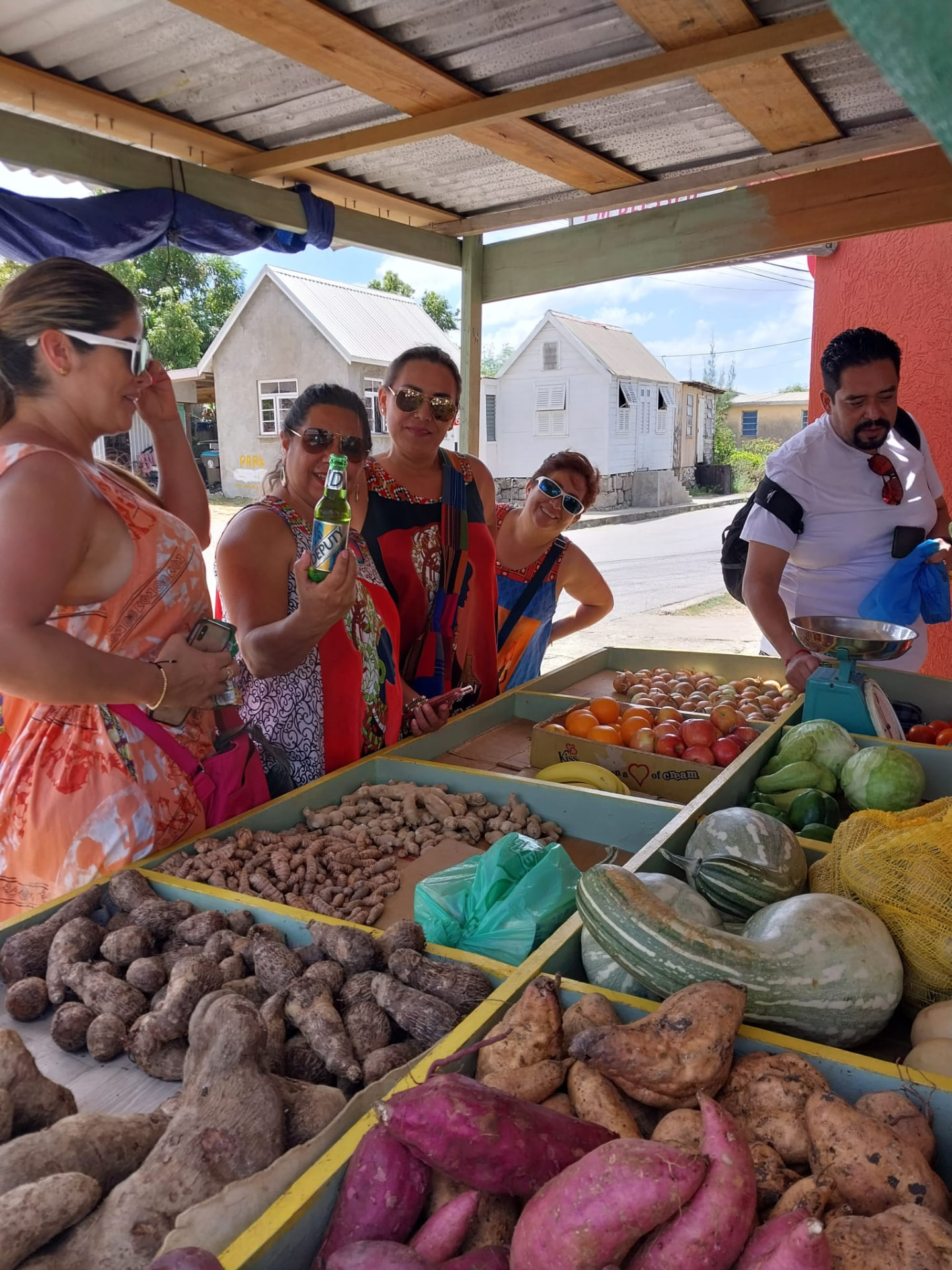 Visitors buying local vegetables at a roadside stall with traditional Bajan chattel houses behind, on an On The House Taxi Service shopping trip